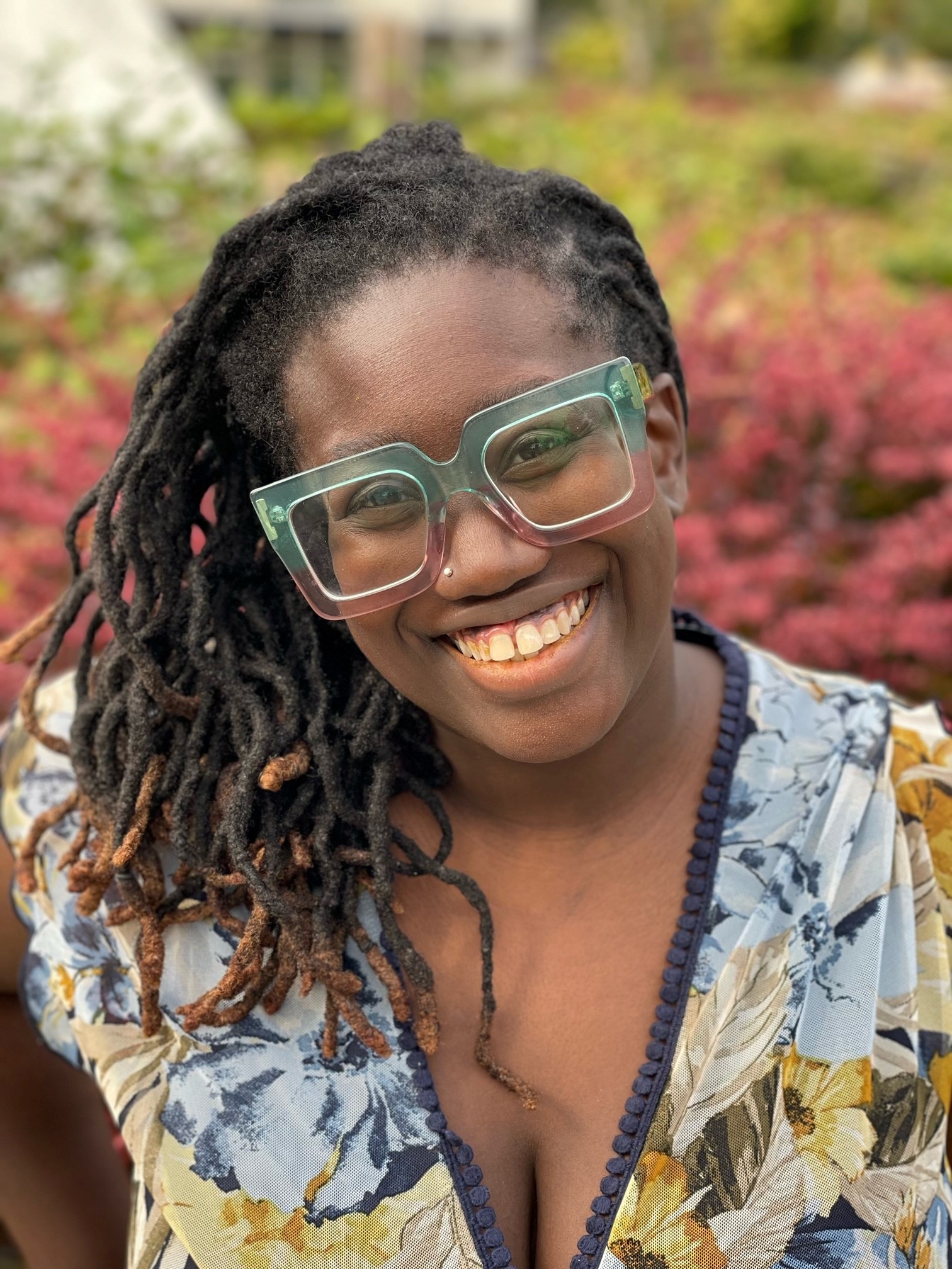 Image of Tolu Taiwo with blue and purple glasses smiling in front of a reddish bush.